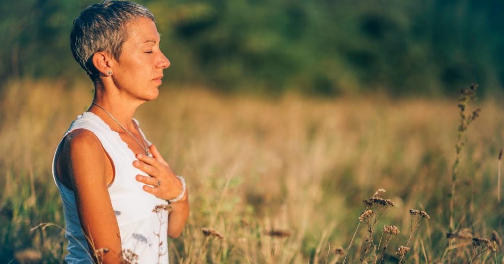 A woman in a field practicing mindfulness with her hand over her heart and eyes closed